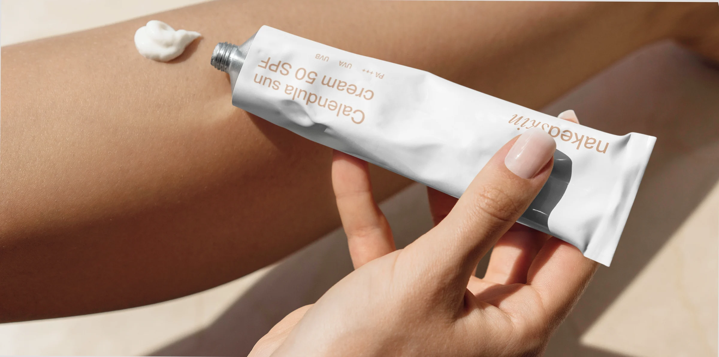 Hand holding a white Calendula SPF 50 sun cream tube beside a dab of cream on a tanned leg, for Naked Skin.