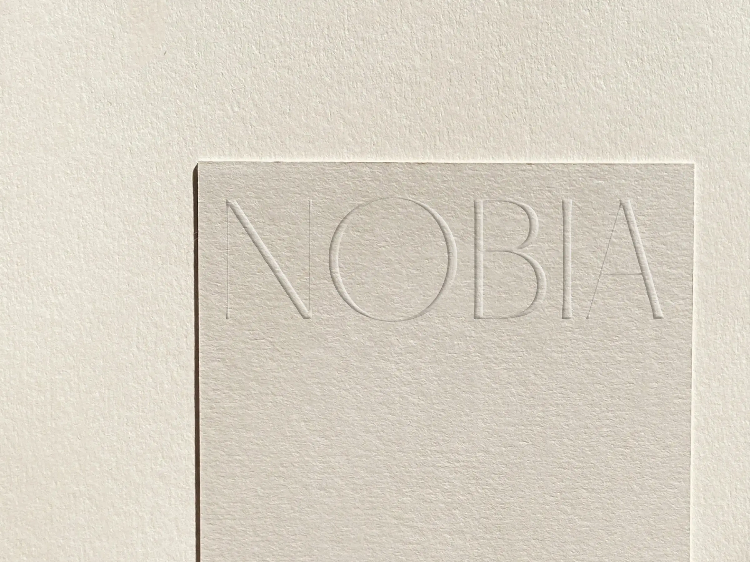 Cream textured paper card with the NOBIA wordmark blind-debossed across the top, on matching paper, for Nobia.